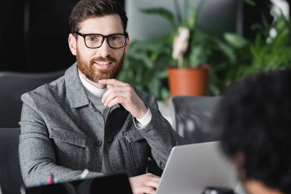 Agente de publicidad positiva en anteojos sonriendo a la cámara cerca de la computadora portátil y colega borrosa - foto de stock