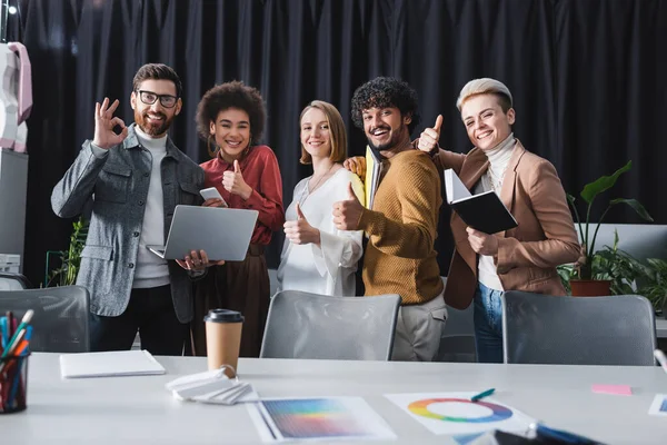 Multiethnic advertising agents smiling at camera while showing thumbs up and okay gestures — Stock Photo