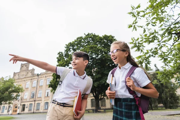 Asiático escolar con cuadernos apuntando con el dedo cerca de compañero de clase al aire libre - foto de stock
