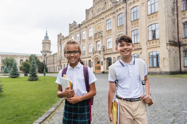 Studenti multietnici positivi con quaderni che guardano la macchina fotografica vicino alla scuola offuscata all'aperto — Foto stock