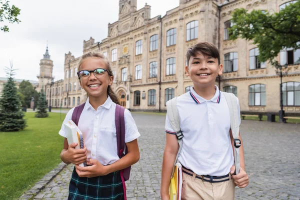 Fröhliche interrassische Schulkinder mit Rucksäcken und Notizbüchern, die draußen spazieren gehen — Stockfoto
