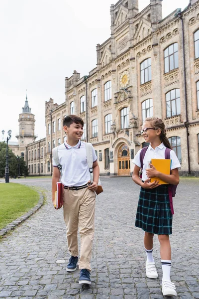Позитивні багатоетнічні школярі розмовляють під час ходьби поблизу школи на відкритому повітрі — стокове фото