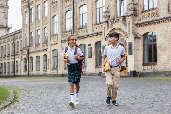 Schoolgirl with notebooks talking to asian friend with backpack while walking near school outdoors — Stock Photo