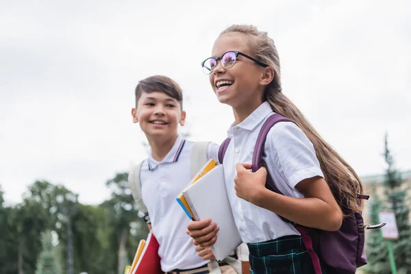 Positive Schulkind mit Rucksack und Notizbüchern zu Fuß in der Nähe verschwommenen asiatischen Freund im Freien — Stockfoto