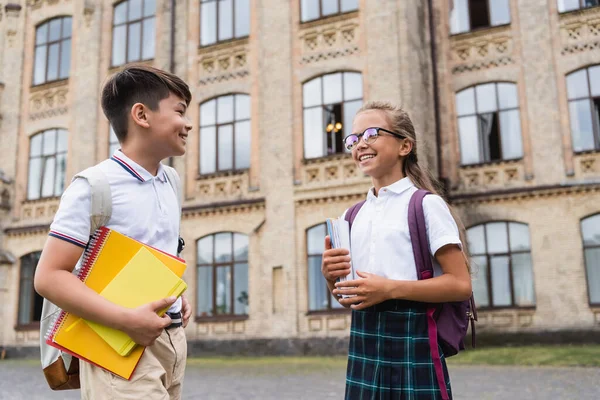 Cheerful interracial pupils with notebooks talking near blurred school outdoors — Stock Photo