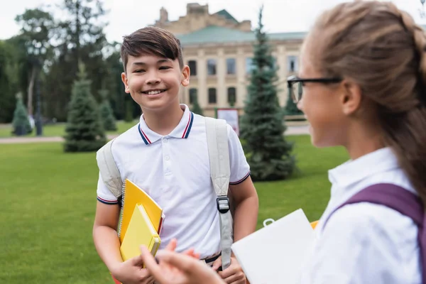 Sonriente asiático escolar celebración de cuadernos cerca borrosa amigo al aire libre - foto de stock