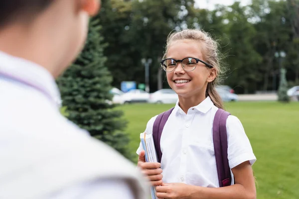 Colegiala positiva en gafas con cuadernos cerca borrosa amigo al aire libre - foto de stock