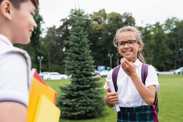 Cheerful pupil in eyeglasses talking to blurred asian friend with notebooks outdoors — Stock Photo