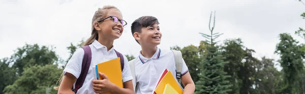 Niños de escuelas multiétnicas sosteniendo cuadernos mientras miran al aire libre, pancarta - foto de stock