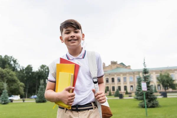 Cheerful asian schoolboy holding copy books and looking at camera outdoors — Stock Photo