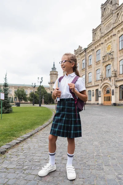 Écolière souriante tenant des cahiers près de l'école floue en plein air — Photo de stock