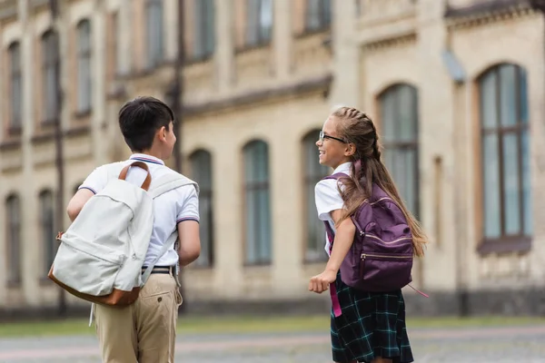 Positive schoolgirl with backpack talking to friend while walking near blurred school — Stock Photo
