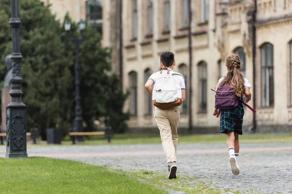 Rückansicht von Vorschulkindern mit Rucksäcken, die in der Nähe der verschwommenen Schule im Freien laufen — Stockfoto