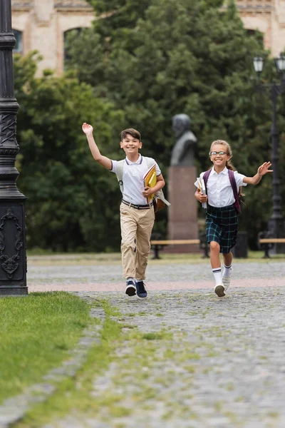 Позитивные межрасовые школьники с блокнотами, машущими руками перед камерой на улице — стоковое фото
