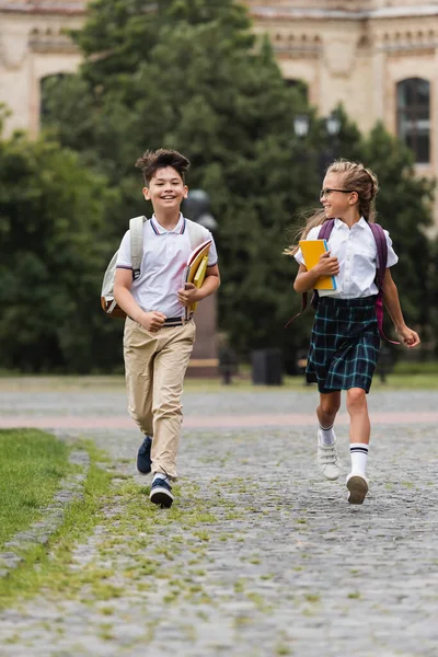 Positive multiethnic pupils with backpacks running on walkway outdoors — Stock Photo