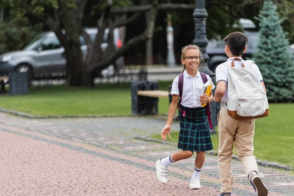 Позитивный школьник в очках разговаривает с другом в рюкзаке на улице — стоковое фото