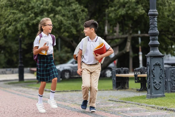 Escolares multiétnicos sonrientes con libros de texto caminando en el parque al aire libre - foto de stock