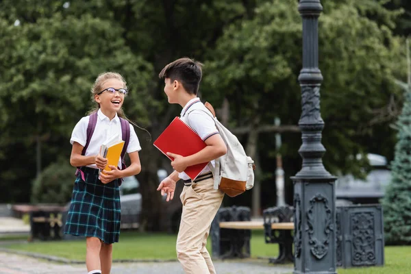 Asian schoolboy with backpack talking to cheerful friend in eyeglasses in park — Stock Photo