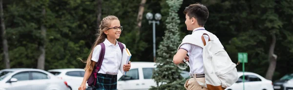 Smiling pupil in eyeglasses holding notebooks near asian friend outdoors, banner — Stock Photo