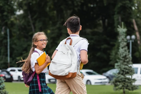 Positive schoolgirl in eyeglasses holding notebooks near classmate outdoors — Stock Photo