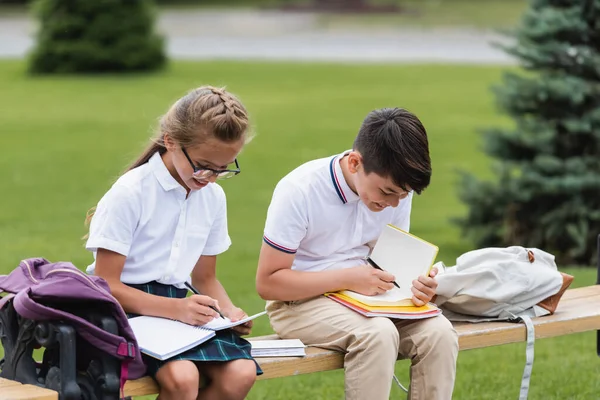 Colegiales multiétnicos escribiendo en cuadernos en el banco al aire libre - foto de stock