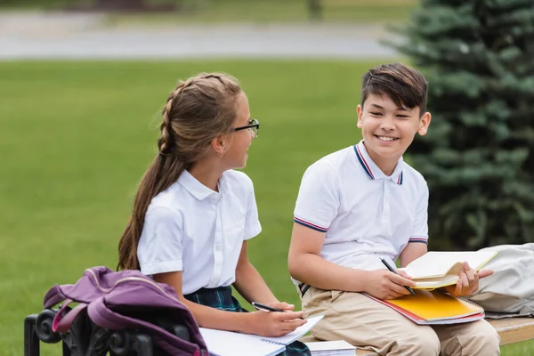 Sonriendo asiático colegial mirando amigo con notebook en banco al aire libre - foto de stock