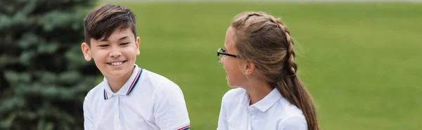 Smiling asian schoolboy looking at friend in eyeglasses outdoors, banner — Stock Photo