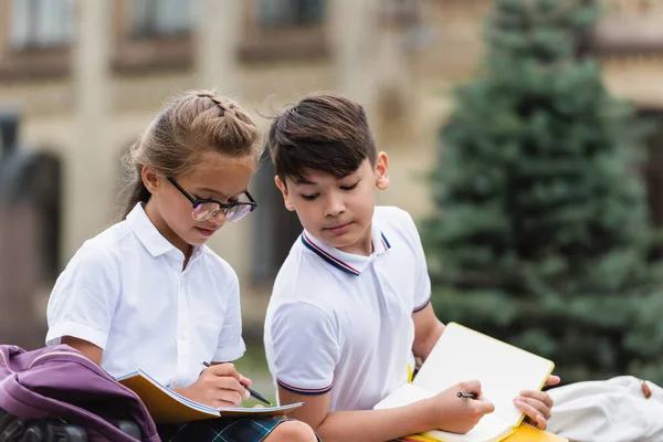 Asian schoolkid looking at notebook near friend with pen outdoors — Stock Photo