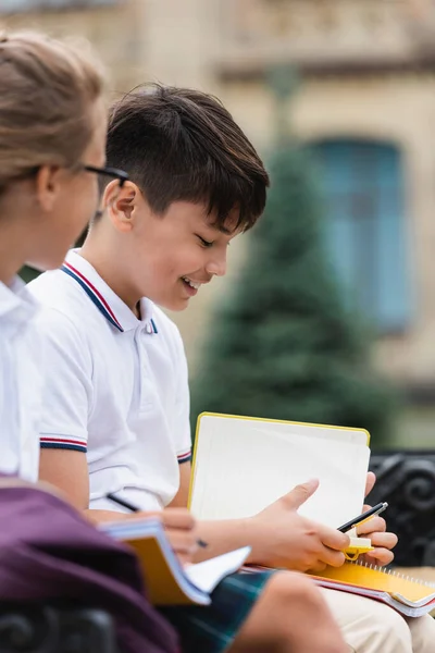 Smiling asian schoolboy holding pen and notebook near blurred friend outdoors — Stock Photo