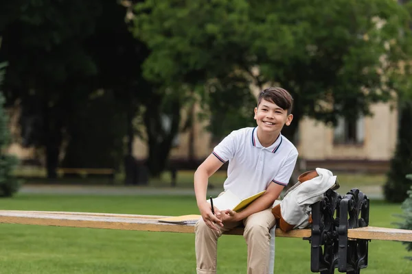 Smiling asian schoolboy holding notebook near backpack on bench outdoors — Stock Photo