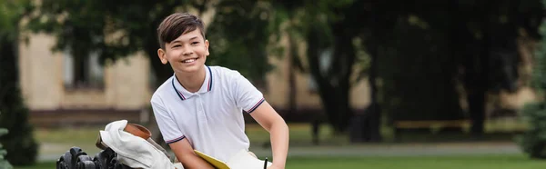 Positive asian schoolboy holding notebook outdoors, banner — Stock Photo