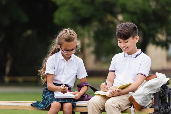 Lächelnde Schüler schreiben auf Notizbücher neben Rucksäcken auf Bank im Freien — Stockfoto