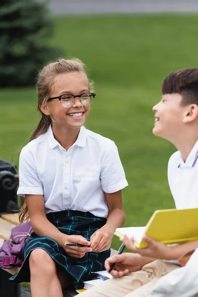 Sonriente colegiala celebración pluma cerca borrosa asiático amigo en banco en parque - foto de stock