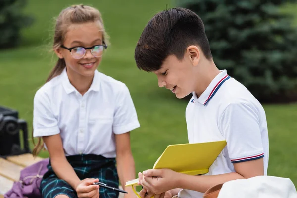 Sonriente asiático escolar celebración notebook cerca amigo con pluma en banco en parque - foto de stock