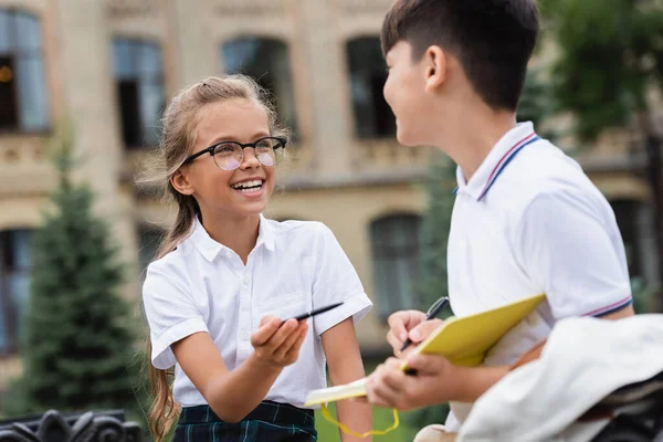 Cheerful schoolgirl with pen looking at asian friend with notebook in park — Stock Photo