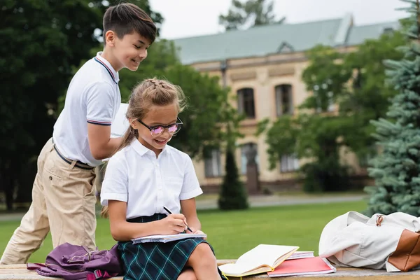 Positives asiatisches Schulkind schaut Freundin beim Schreiben auf Notizbuch auf Bank im Park an — Stockfoto