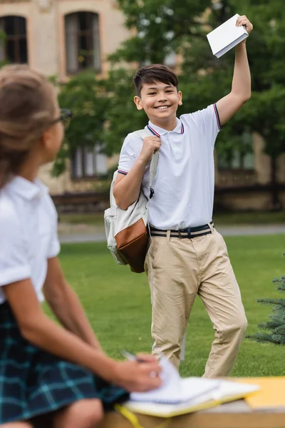 Cheerful asian schoolboy with book and backpack near blurred friend outdoors — Stock Photo