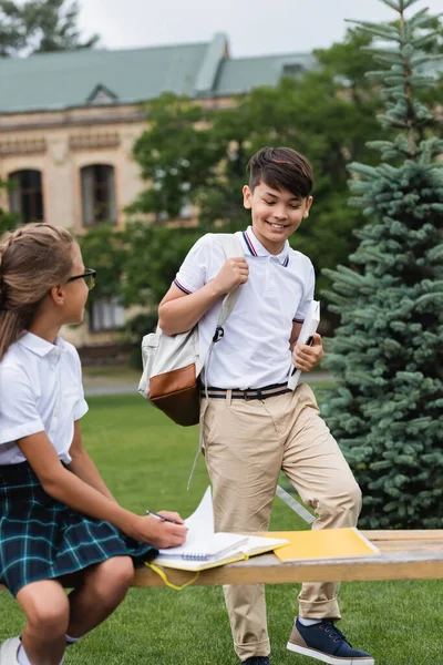 Asian schoolboy holding book near friend on bench in park — Stock Photo