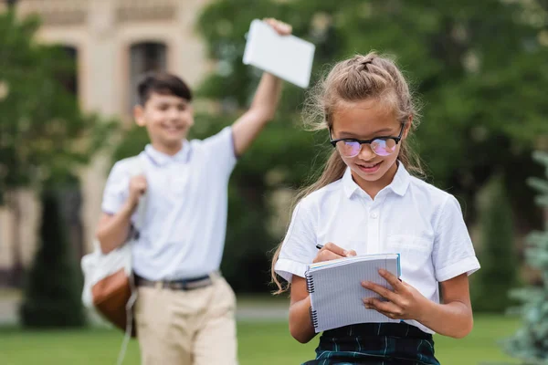 Colegiala escribiendo en el cuaderno cerca borrosa amigo al aire libre - foto de stock