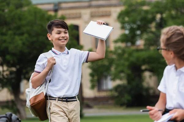 Азійський школяр тримає книжку біля розмитого однокласника в парку. — стокове фото