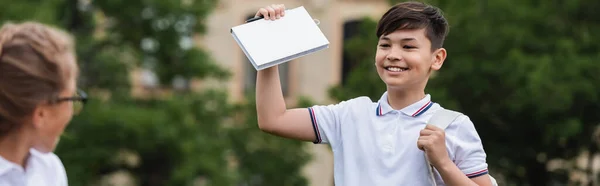 Lächelndes asiatisches Schulkind hält Buch neben verschwommenem Freund im Park, Banner — Stockfoto