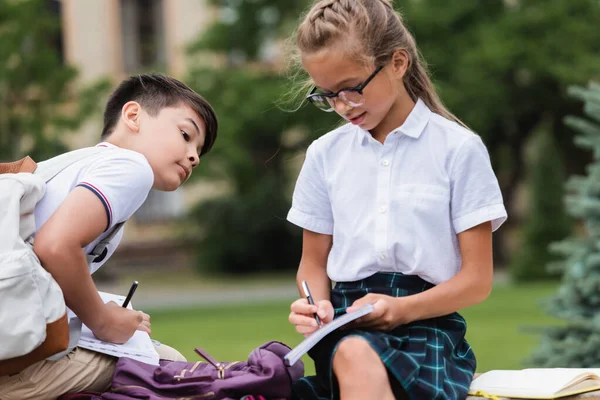 Allievi multietnici che scrivono su quaderni su panchina nel parco — Foto stock