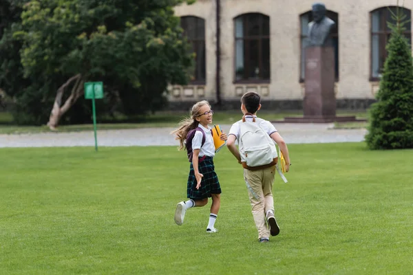 Happy schoolgirl holding notebooks while walking near friend on lawn — Stock Photo