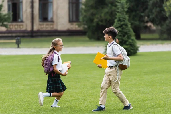 Улыбающиеся одноклассники с блокнотами, бегающими по газону в парке — стоковое фото