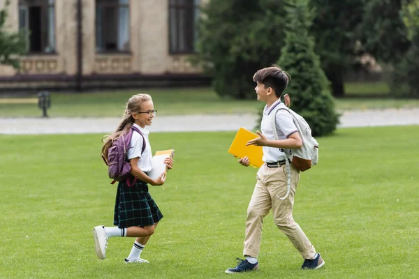 Smiling multiethnic classmates with notebooks running on lawn in park — Stock Photo