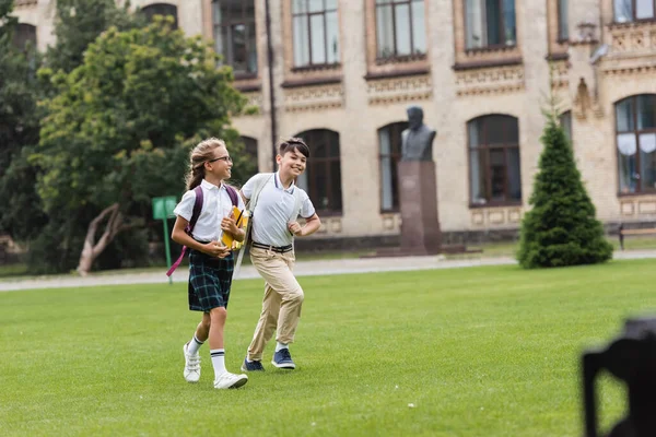 Позитивные межрасовые одноклассники с блокнотами, гуляющими по траве в парке — стоковое фото
