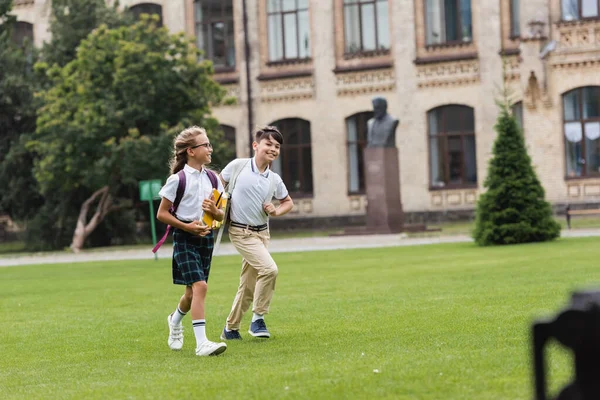 Des camarades de classe interraciaux positifs avec des cahiers marchant sur l'herbe dans le parc — Photo de stock