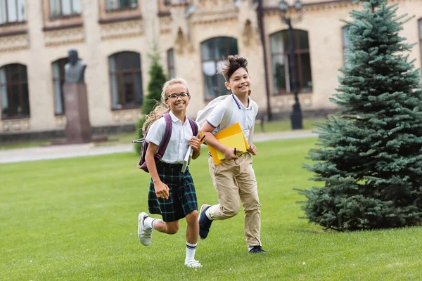 Smiling multiethnic schoolkids with notebooks running on lawn in park — Stock Photo