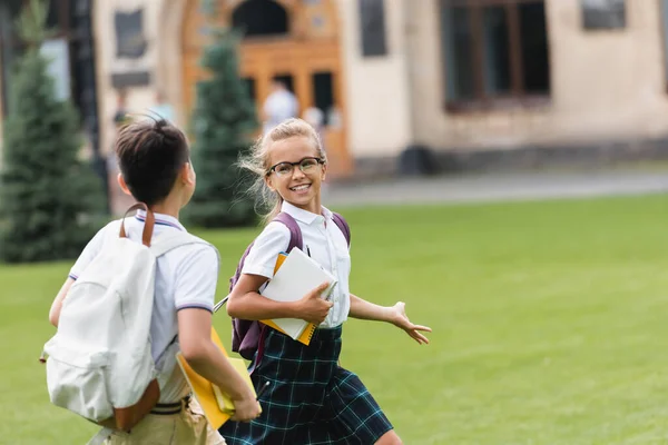 Studentessa sorridente con quaderni che cammina vicino al compagno di classe sul prato nel parco — Foto stock