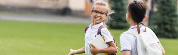 Positive schoolgirl with notebooks walking near friend on lawn in park, banner — Stock Photo